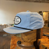 The Shop hat by Ballad of the Bird Dog features a light blue design and a white-and-black oval patch reading "Ballad of the Bird Dog, Richmond, TX," shown on a wooden form in a rustic setting.