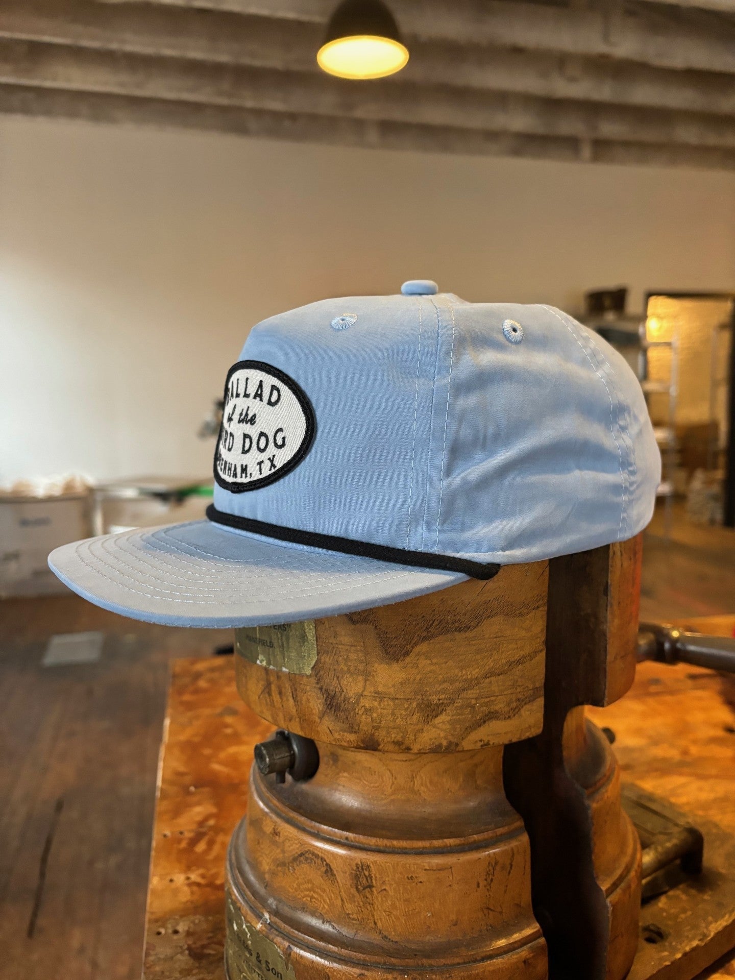 The Shop hat by Ballad of the Bird Dog features a light blue design and a white-and-black oval patch reading "Ballad of the Bird Dog, Richmond, TX," shown on a wooden form in a rustic setting.