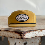 The Shop Hat | Classic Original Oval Logo by Ballad of the Bird Dog features a mustard yellow flat-brim design and a signature oval patch embroidered with “Ballad of the Bird Dog, Brenham, TX.” Resting on a rustic metal surface, it stands out as a best seller from the Ballad Gear collection.