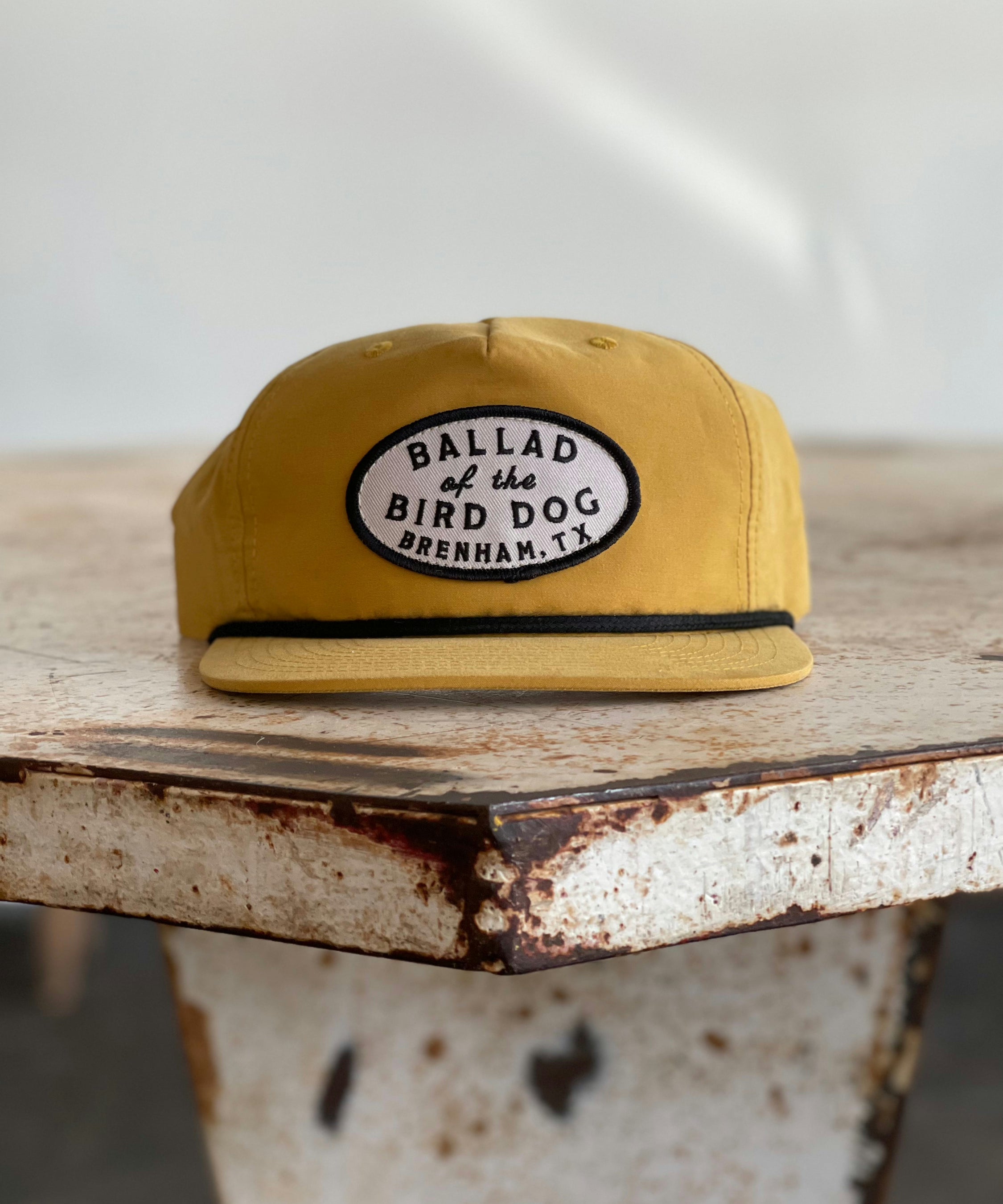 The Shop Hat | Classic Original Oval Logo by Ballad of the Bird Dog features a mustard yellow flat-brim design and a signature oval patch embroidered with “Ballad of the Bird Dog, Brenham, TX.” Resting on a rustic metal surface, it stands out as a best seller from the Ballad Gear collection.