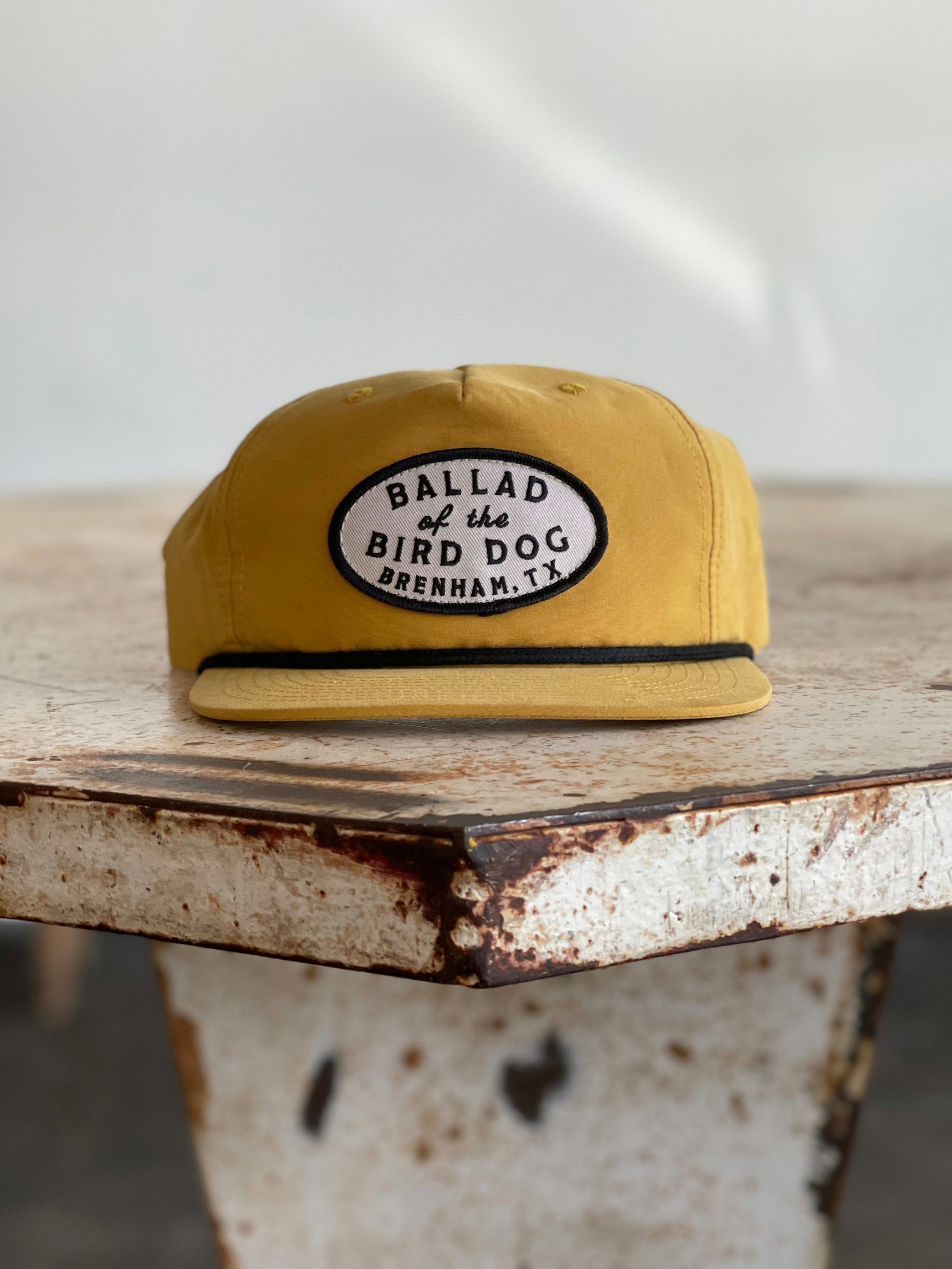 The Shop Hat | Classic Original Oval Logo by Ballad of the Bird Dog features a mustard yellow flat-brim design and a signature oval patch embroidered with “Ballad of the Bird Dog, Brenham, TX.” Resting on a rustic metal surface, it stands out as a best seller from the Ballad Gear collection.