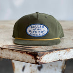 The Shop Hat | Classic Original Oval Logo from Ballad of the Bird Dog, featuring an olive green design and a blue and white patch reading “Ballad of the Bird Dog Brenham, TX,” rests on a weathered, rusty metal table—one of the top-selling hats from the Ballad Gear collection.
