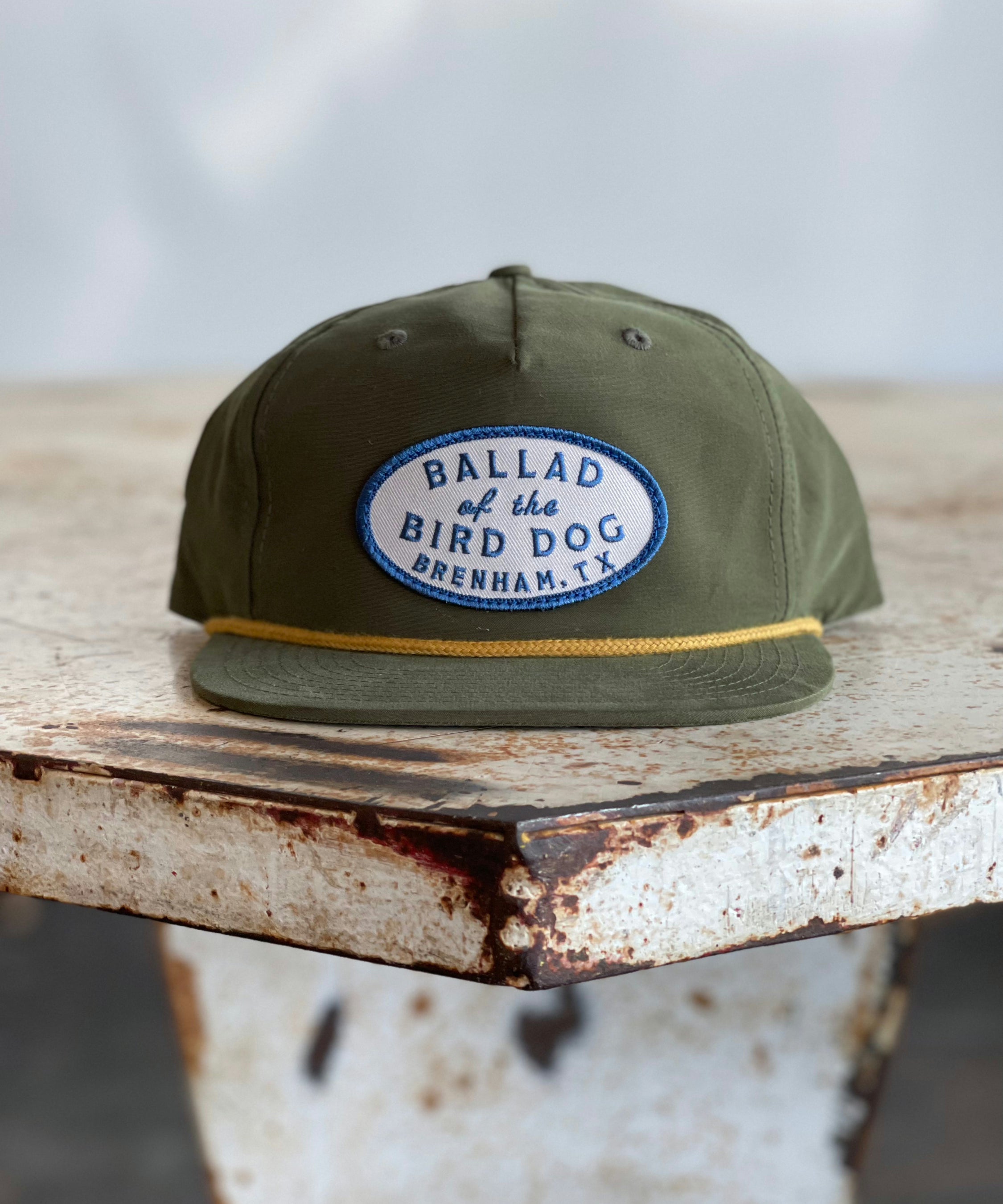 The Shop Hat | Classic Original Oval Logo from Ballad of the Bird Dog, featuring an olive green design and a blue and white patch reading “Ballad of the Bird Dog Brenham, TX,” rests on a weathered, rusty metal table—one of the top-selling hats from the Ballad Gear collection.