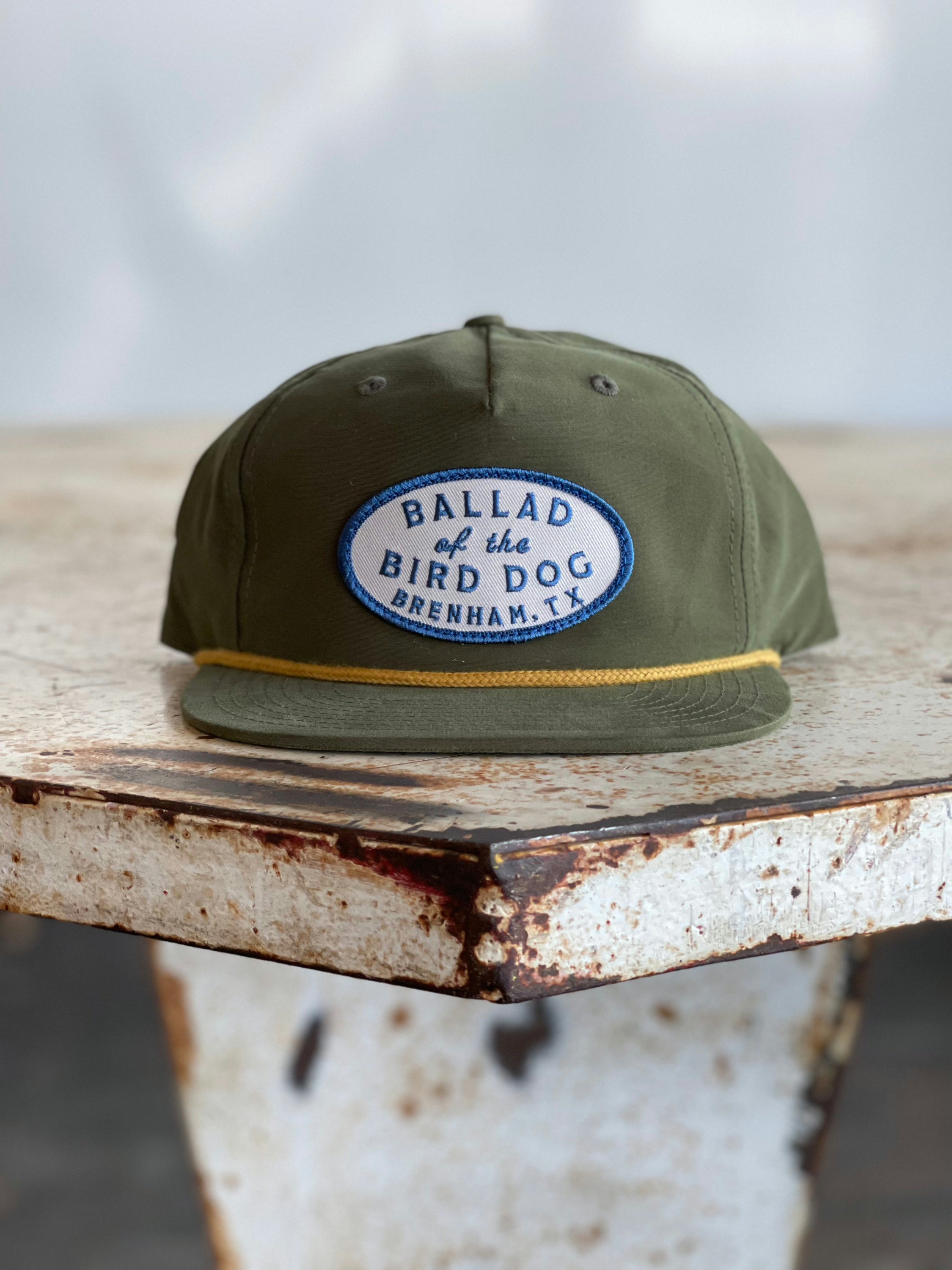 The Shop Hat | Classic Original Oval Logo from Ballad of the Bird Dog, featuring an olive green design and a blue and white patch reading “Ballad of the Bird Dog Brenham, TX,” rests on a weathered, rusty metal table—one of the top-selling hats from the Ballad Gear collection.