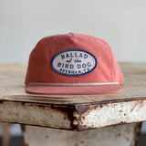 A Shop Hat from Ballad of the Bird Dog, featuring their classic original oval logo and a patch reading “Ballad of the Bird Dog Brenham, TX,”—one of our best sellers—rests on a weathered wooden table.
