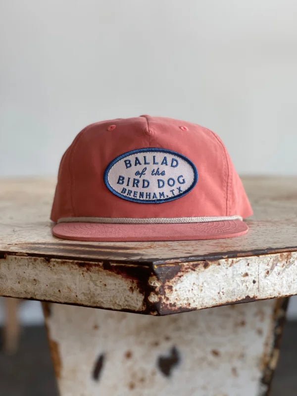 A Shop Hat from Ballad of the Bird Dog, featuring their classic original oval logo and a patch reading “Ballad of the Bird Dog Brenham, TX,”—one of our best sellers—rests on a weathered wooden table.