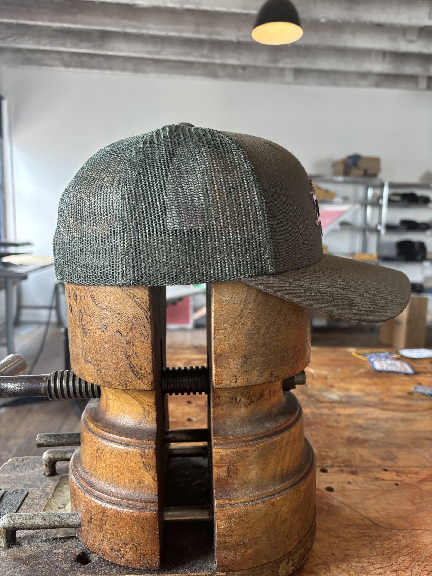 The Shop Hat | Desert Dog by Ballad of the Bird Dog, a rugged green mesh cap, is held sideways in a wooden vice on a workbench in a workshop with shelves and boxes in the background.