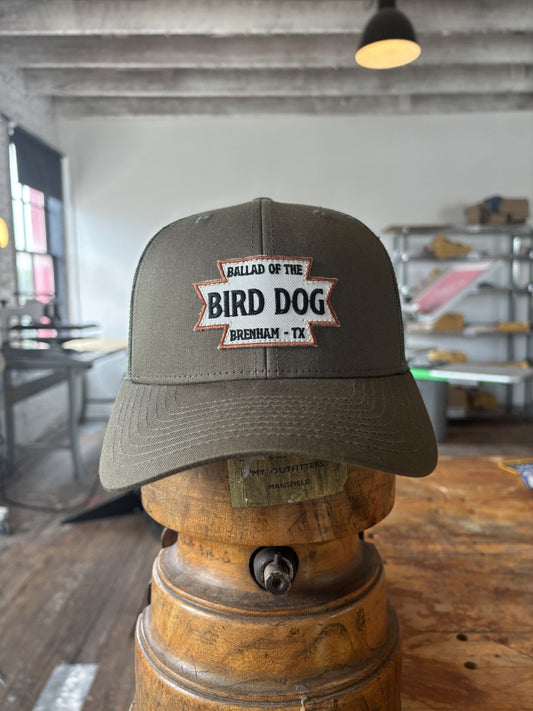 The Shop Hat | Desert Dog by Ballad of the Bird Dog, featuring a "Ballad of the Bird Dog Brenham, TX" patch, sits on a wooden stand in a rustic indoor setting—a rugged accessory with desert-inspired flair.