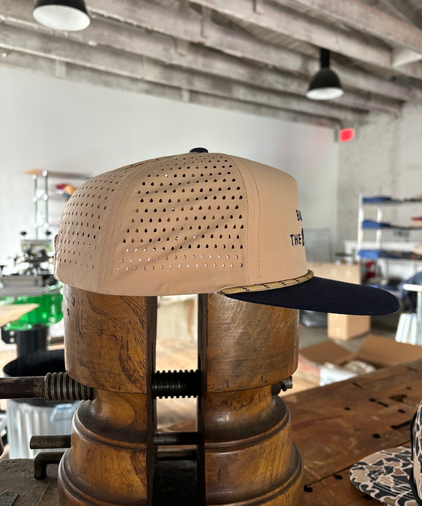 The Ballad of the Bird Dog Shop Hat from the Desert Series, featuring tan and navy colors with perforated sides, is displayed on a vintage hat stretcher in a workshop setting.
