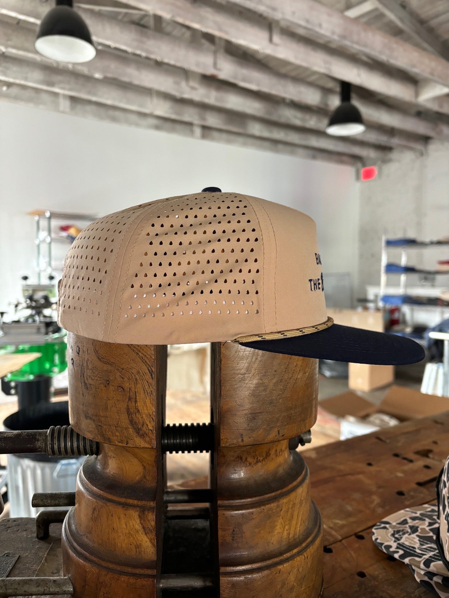 The Ballad of the Bird Dog Shop Hat from the Desert Series, featuring tan and navy colors with perforated sides, is displayed on a vintage hat stretcher in a workshop setting.
