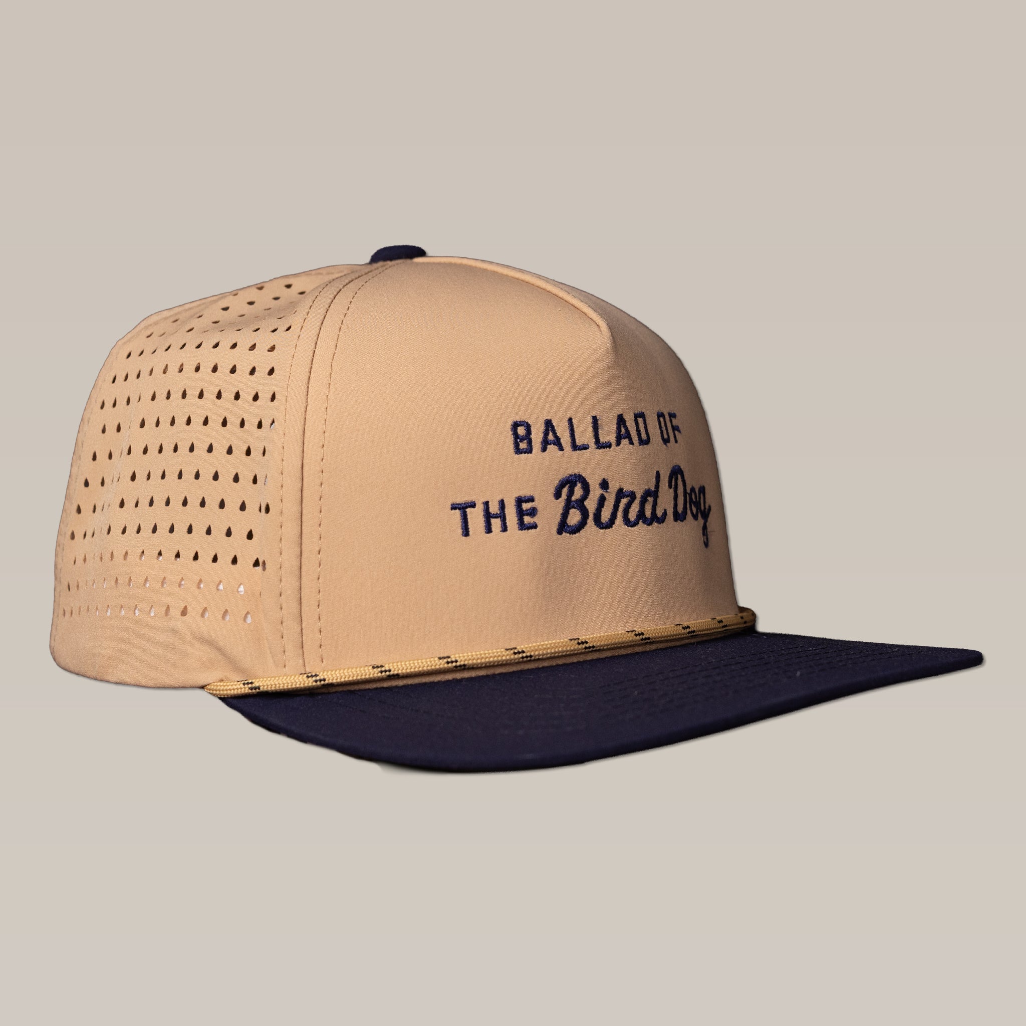 The Ballad of the Bird Dog Shop Hat from the Desert Series features tan and navy colors, "Ballad of the Bird Dog" text on the front, and breathable perforated side panels, shown against a neutral background.