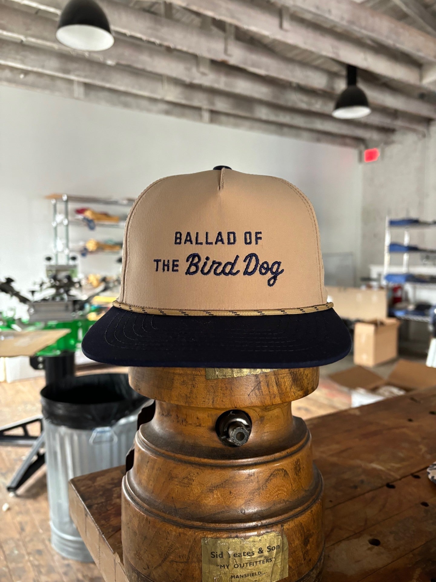 The Ballad of the Bird Dog Shop Hat | Desert Series, a beige and black cap, rests on a wooden object in a workshop or studio setting.