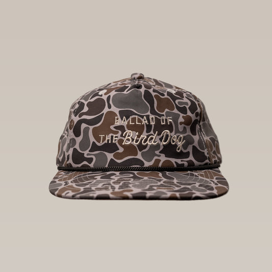 The Ballad of the Bird Dog Shop Hat from the Desert Series features a camouflage pattern and “BALLAD OF THE Bird Dog” embroidery on the front, shown against a plain beige background.