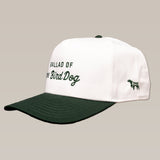 The Shop Hat from the Desert Series by Ballad of the Bird Dog is a white and dark green baseball cap with "Ballad of the Bird Dog" embroidered on the front and a small dog outline on the side.