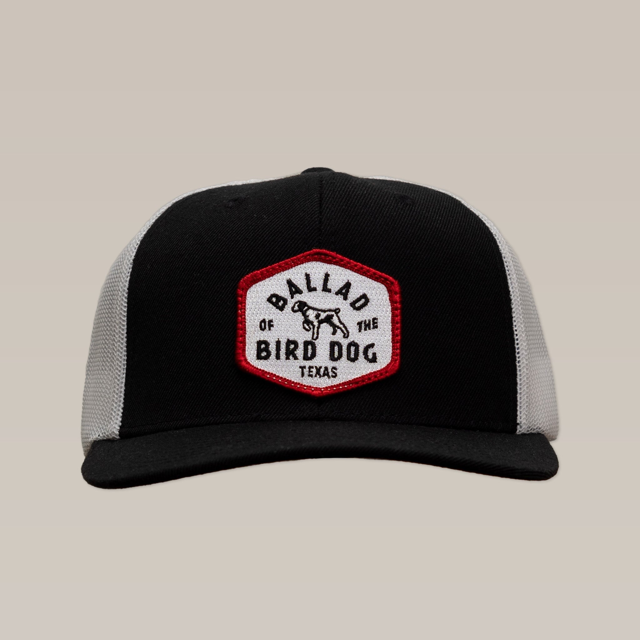 The Field Companion Shop Hat by Ballad of the Bird Dog is a black and gray hat featuring a hexagonal patch with a bird dog graphic and “Ballad of the Bird Dog Texas” text—an original, classic design.