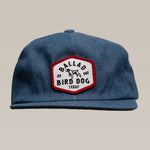 The Ballad of the Bird Dog Shop Hat | Field Companion features a blue cap with a red and white hexagonal patch reading “Ballad of the Bird Dog Texas” and a dog image—a heritage-inspired accessory for symbolic headwear fans.