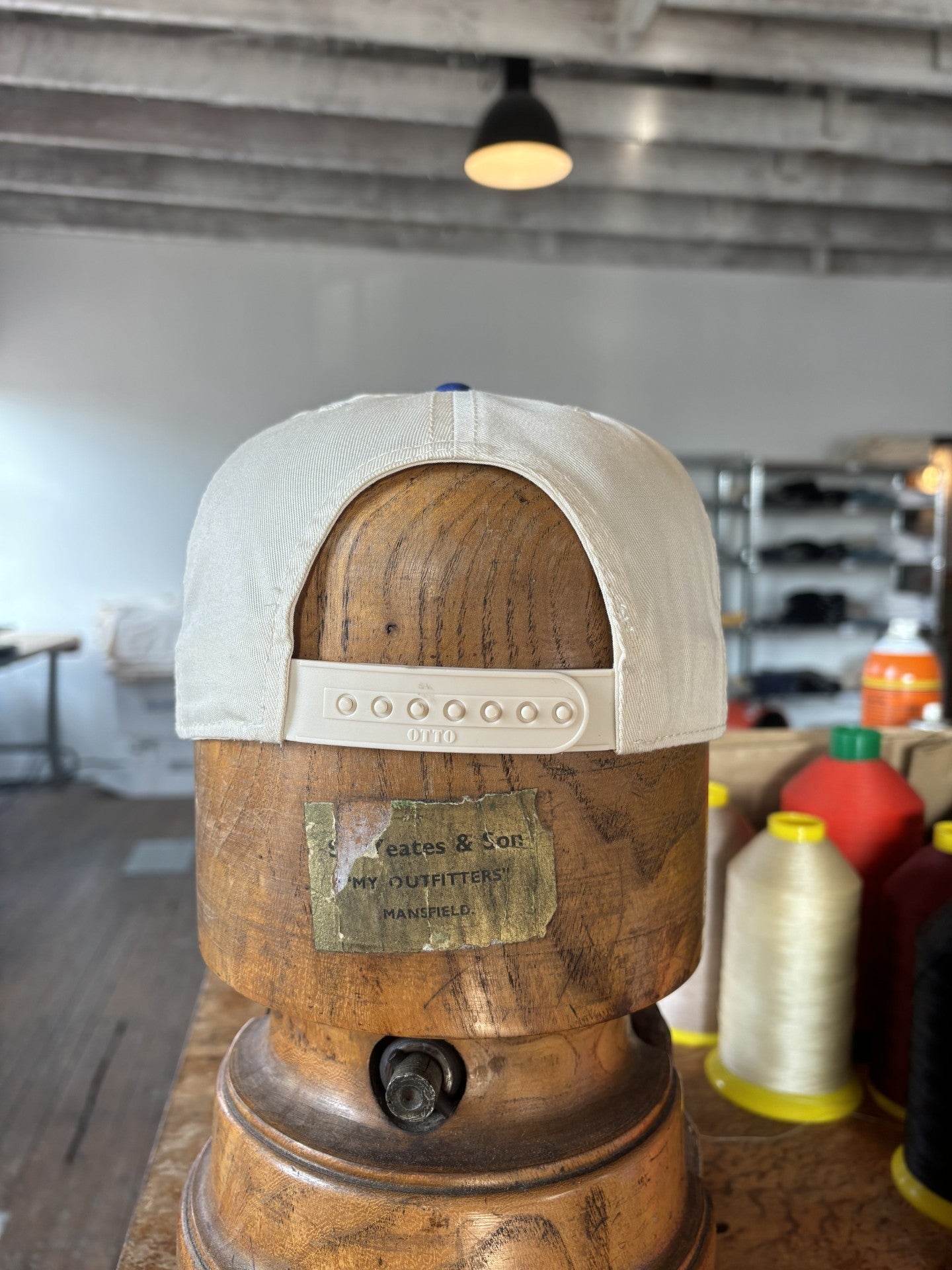 The Shop Hat | Field Companion by Ballad of the Bird Dog, a white adjustable baseball cap, is displayed on a wooden hat stand with spools of thread and shelves in the background.