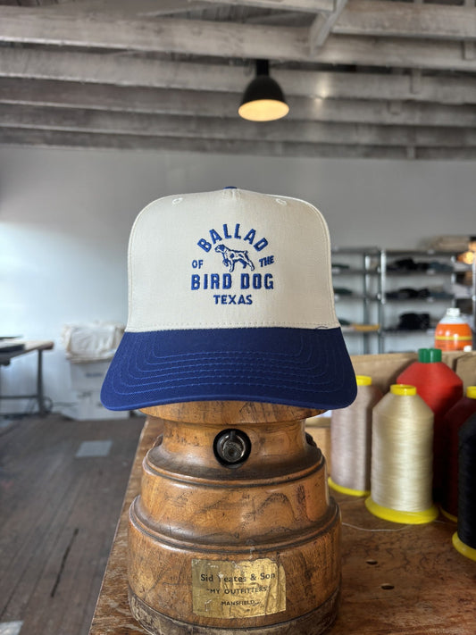 The Ballad of the Bird Dog Shop Hat | Field Companion baseball cap is showcased on a wooden stand in a workshop, with spools of thread in the background.