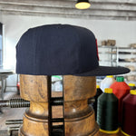 The navy blue Shop Hat | Field Companion by Ballad of the Bird Dog is shown on a wooden hat form in a workspace, surrounded by sewing thread spools and shelves in the background.