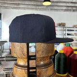 The Shop Hat | Field Companion by Ballad of the Bird Dog—a navy heritage hat—rests on a vintage wooden stretcher amid spools of thread and workshop shelves in the background.