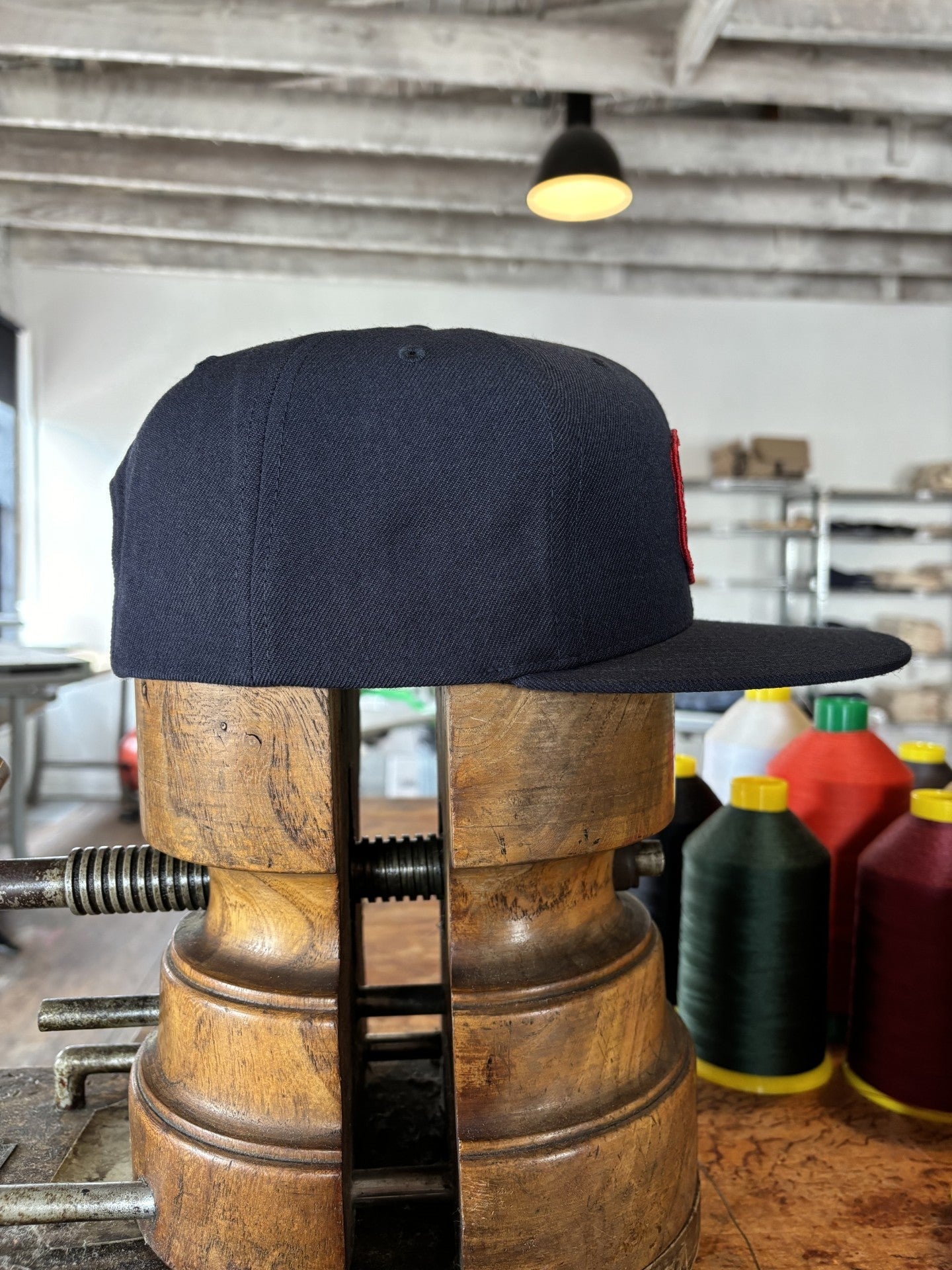 The Shop Hat | Field Companion by Ballad of the Bird Dog—a navy heritage hat—rests on a vintage wooden stretcher amid spools of thread and workshop shelves in the background.