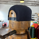 The Ballad of the Bird Dog Shop Hat | Field Companion is a black snapback cap, shown on a wooden hat block with sewing threads and equipment in the background.