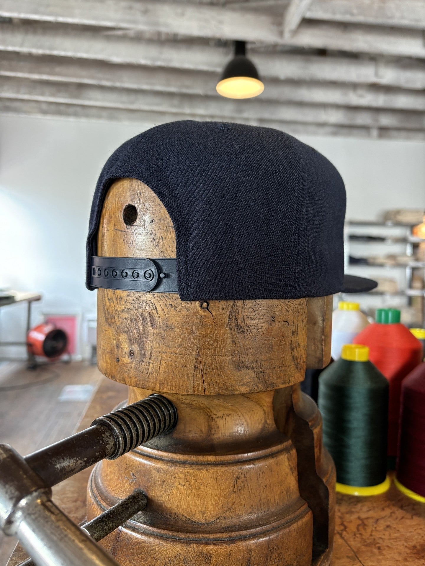 The Ballad of the Bird Dog Shop Hat | Field Companion is a black snapback cap, shown on a wooden hat block with sewing threads and equipment in the background.