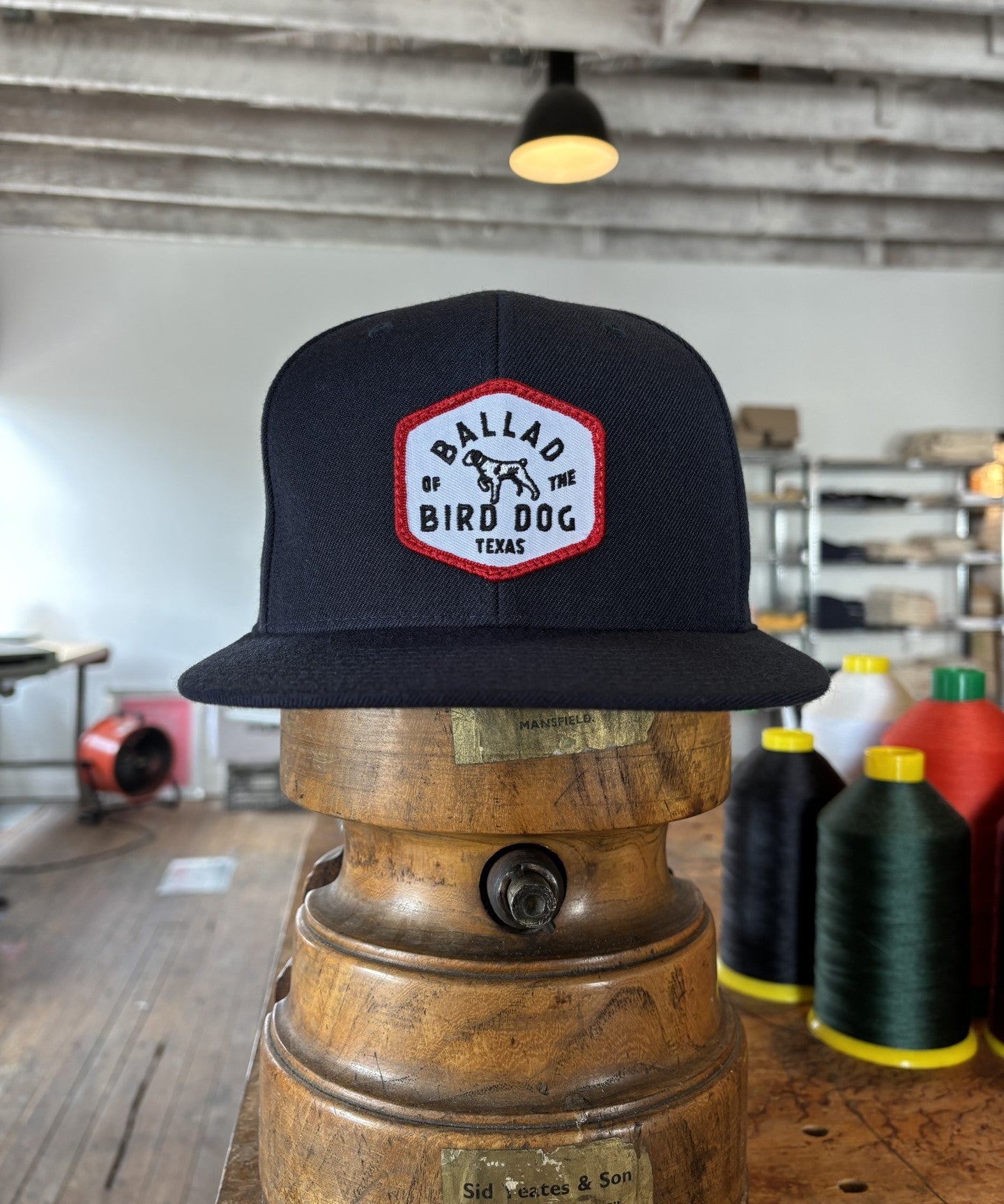 The Ballad of the Bird Dog Shop Hat | Field Companion, featuring a red and white “Ballad of the Bird Dog Texas” patch, is displayed on a vintage wooden stand in a workshop amid sewing supplies.