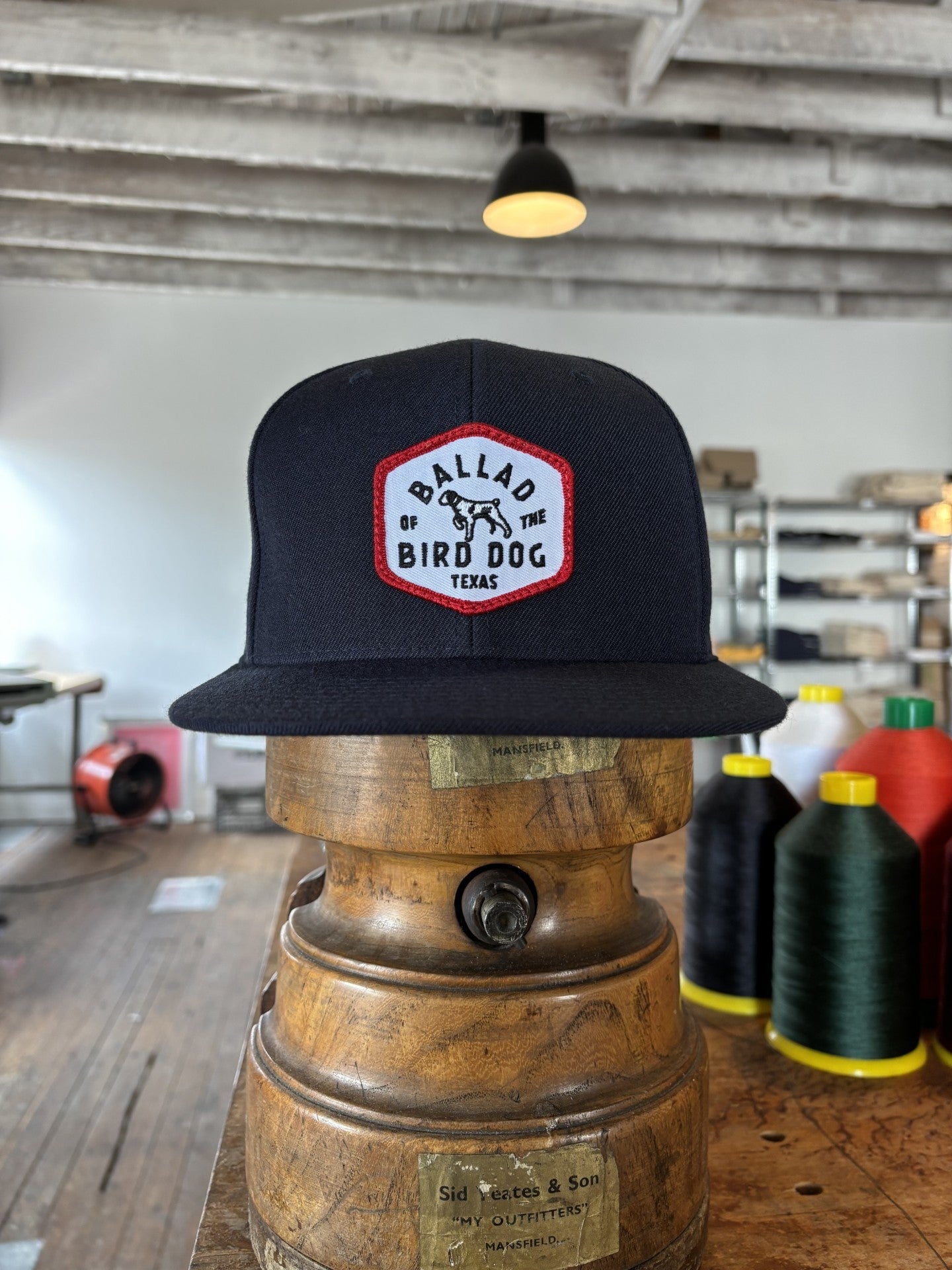 The Ballad of the Bird Dog Shop Hat | Field Companion, featuring a red and white “Ballad of the Bird Dog Texas” patch, is displayed on a vintage wooden stand in a workshop amid sewing supplies.