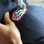 A close-up of a sewing machine stitching a Shop Hat | Field Companion by Ballad of the Bird Dog, with a person's hand holding the navy blue, heritage-inspired hat in place.