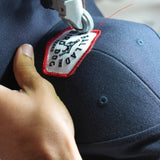 A close-up of a sewing machine stitching a Field Companion patch onto the Shop Hat by Ballad of the Bird Dog, creating a unique navy blue cap held in hand.