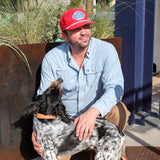 A man wearing a Shop Hat | Home Base by Ballad of the Bird Dog and a blue shirt kneels outside, petting a black and white dog. A mural and plants can be seen in the background.