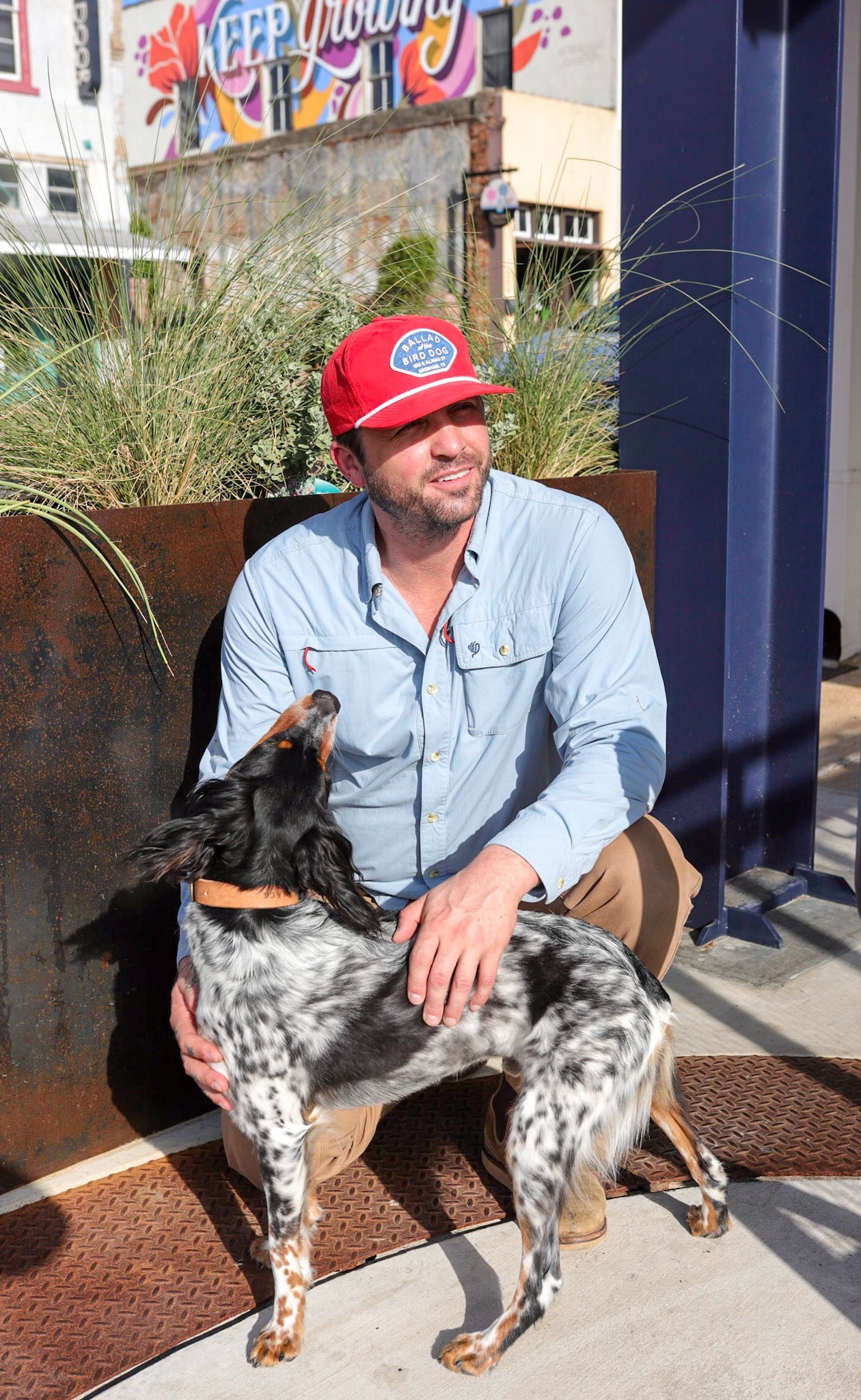 A man wearing a Shop Hat | Home Base by Ballad of the Bird Dog and a blue shirt kneels outside, petting a black and white dog. A mural and plants can be seen in the background.