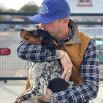A person in a blue Shop Hat | Home Base by Ballad of the Bird Dog and a plaid shirt sits outdoors, hugging and kissing a black and white dog.