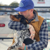 A man wearing the Ballad of the Bird Dog Shop Hat | Home Base hugs and kisses a black and white dog while sitting outdoors in front of a metal fence.