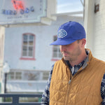 A man in a tan quilted vest, plaid shirt, and Ballad of the Bird Dog's Shop Hat | Home Base stands outdoors near a white building with blurred signage, wearing community headwear that supports small business.