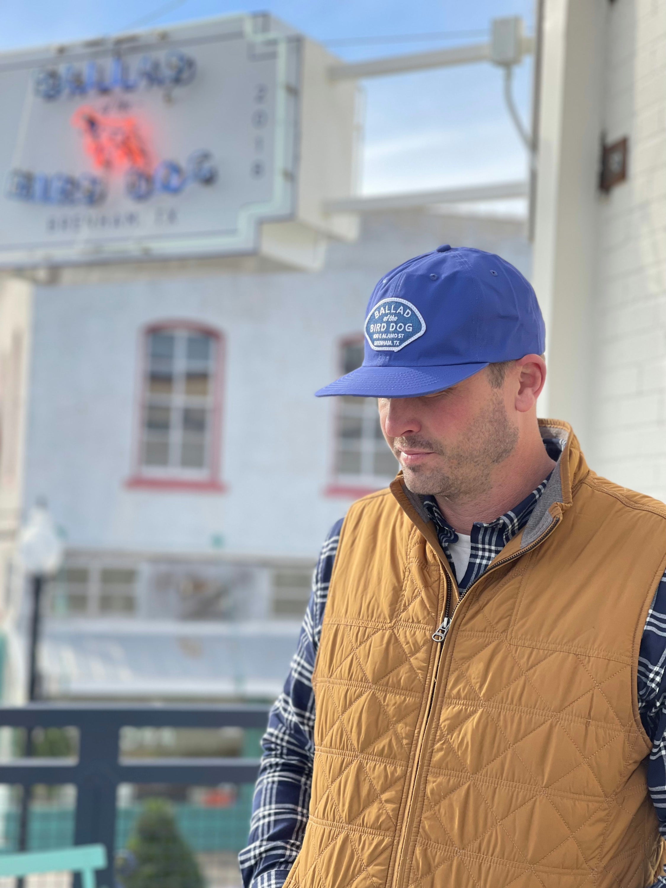 A man wears the Shop Hat | Home Base by Ballad of the Bird Dog, paired with a plaid shirt and tan vest, standing outside with a blurred building and sign in the background.