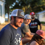 Four people sit outdoors in sunlight, wearing casual clothes and hats—including the Shop Hat | Home Base by Ballad of the Bird Dog. Trees and buildings are behind them, with a smiling man in branded headwear at the front.