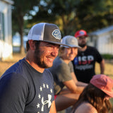 A man in a blue Under Armour shirt and cap smiles while sitting outdoors with three others, all in casual clothes and wearing hats like the Shop Hat | Home Base by Ballad of the Bird Dog. Trees and buildings create a relaxed community vibe.