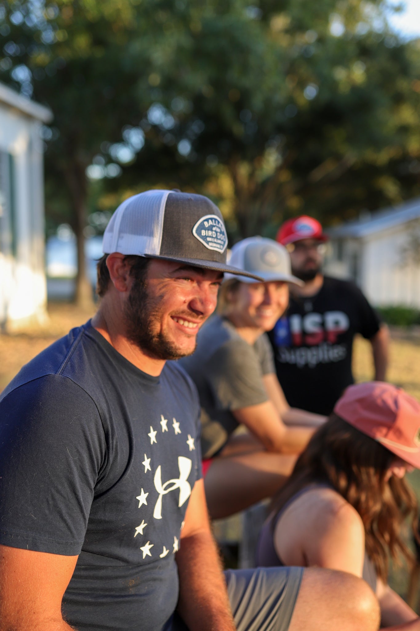 A man in a blue Under Armour shirt and cap smiles while sitting outdoors with three others, all in casual clothes and wearing hats like the Shop Hat | Home Base by Ballad of the Bird Dog. Trees and buildings create a relaxed community vibe.