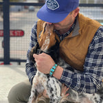 A man in a blue Shop Hat | Home Base by Ballad of the Bird Dog and a brown vest kneels, hugging a dog that licks his face.