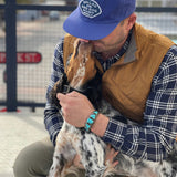 A man in a Shop Hat | Home Base by Ballad of the Bird Dog and a brown vest kneels to hug a dog that's licking his face.