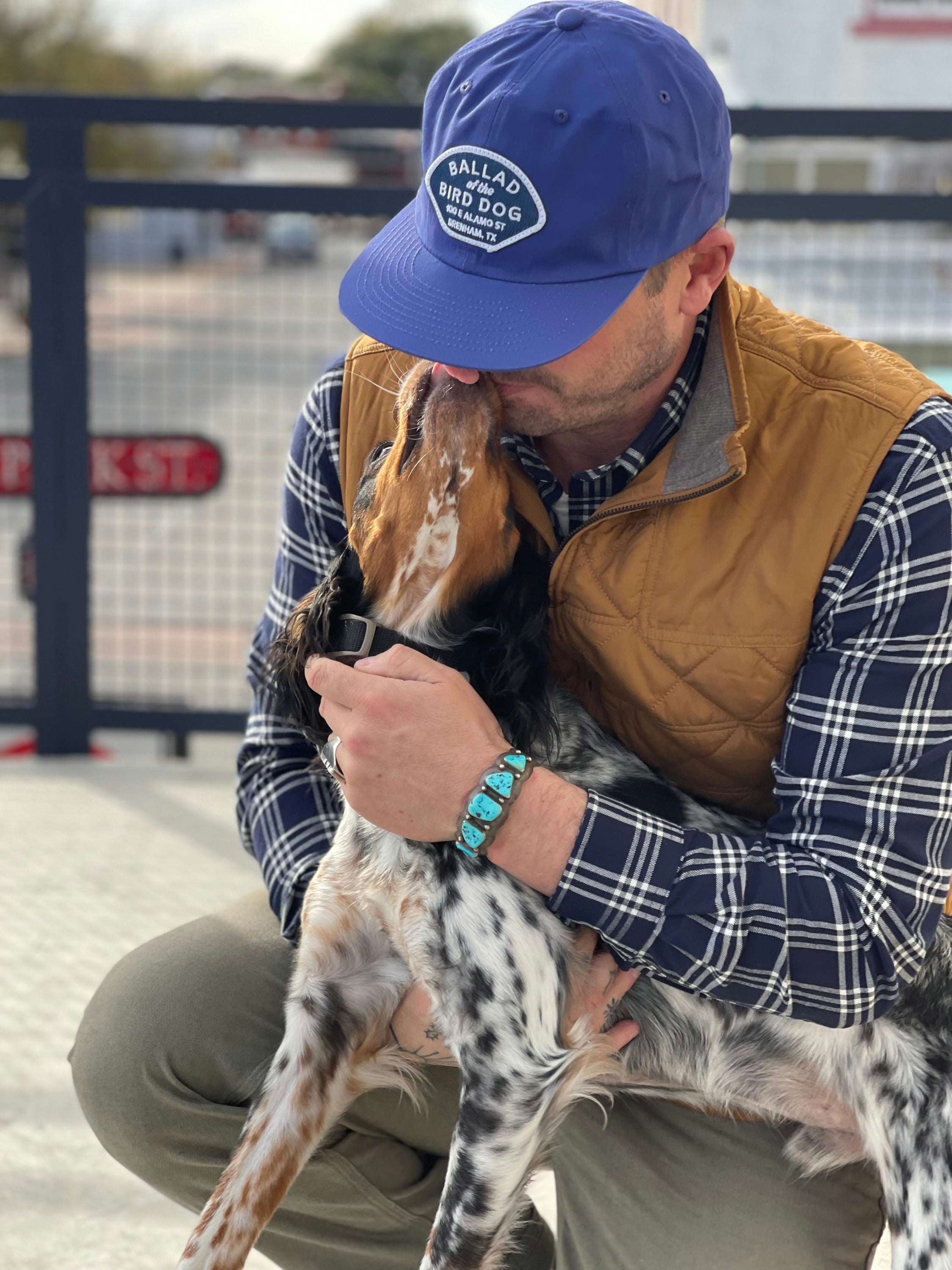 A man in a Shop Hat | Home Base by Ballad of the Bird Dog and a brown vest kneels to hug a dog that's licking his face.
