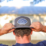 A person adjusts a gray Shop Hat | Home Base by Ballad of the Bird Dog outdoors, highlighting this branded community headwear with a blurred building and trees in the background.