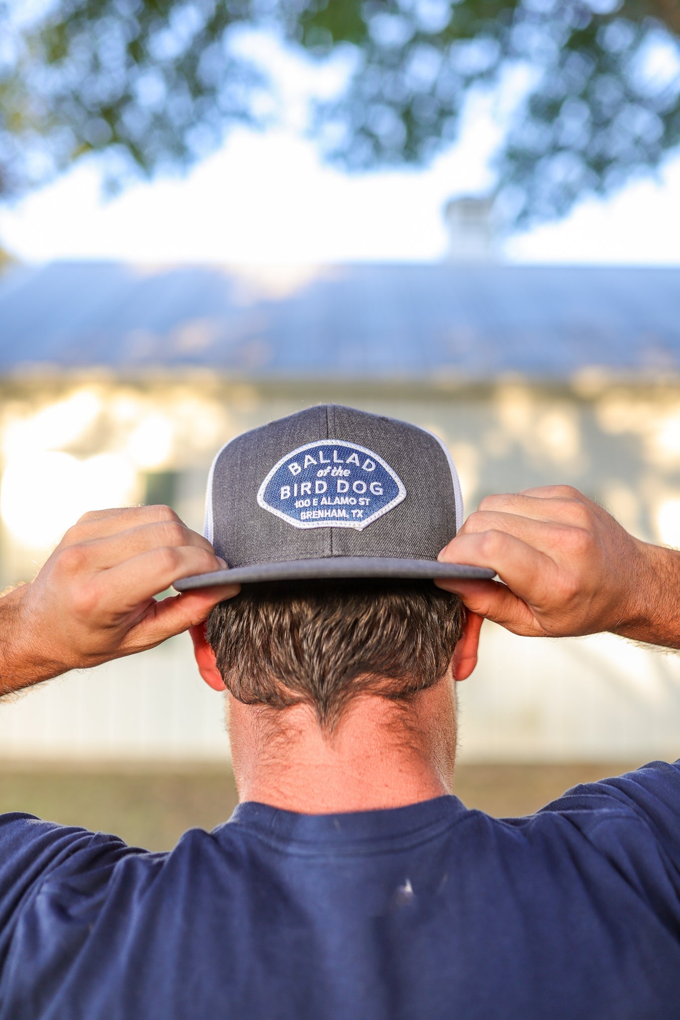 A man, seen from behind, adjusts a Shop Hat | Home Base by Ballad of the Bird Dog while standing outside near a light-colored building.