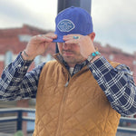 A person wearing a blue "Shop Hat | Home Base | Ballad of the Bird Dog" by Ballad of the Bird Dog, along with a plaid shirt and brown quilted vest, stands outdoors adjusting the hat’s brim with both hands.