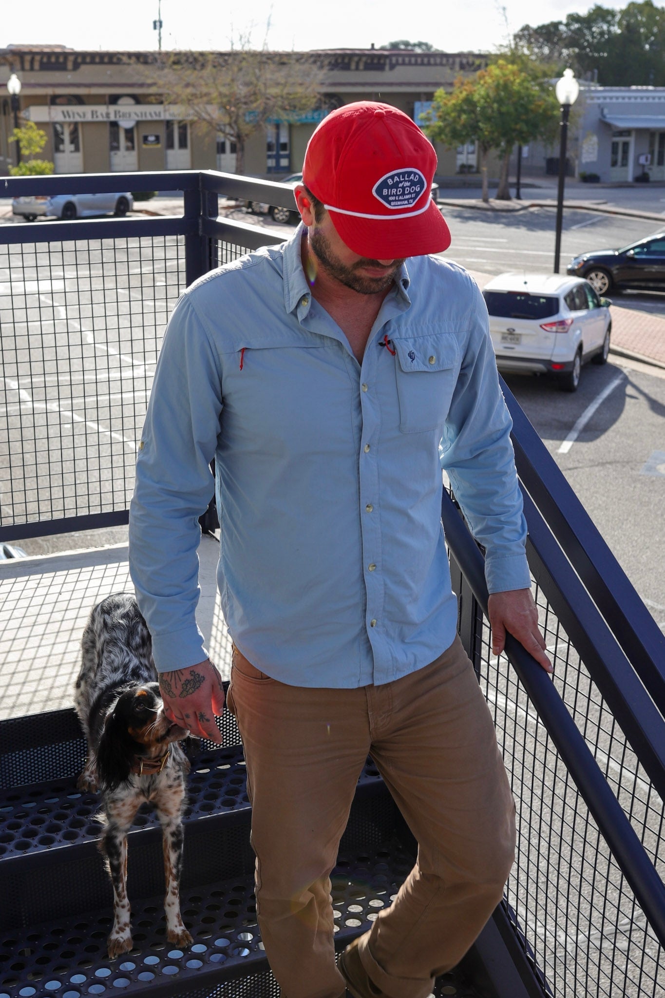 A man in a Ballad of the Bird Dog Shop Hat (Home Base) and a light blue shirt walks up an outdoor metal staircase with a black and white dog beside him.