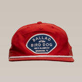 The Shop Hat | Home Base by Ballad of the Bird Dog is a red cap featuring a blue and white patch reading "Ballad of the Bird Dog, 100 E Alamo St, Brenham, TX"—a great way to support a local small business and community.