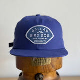 The Shop Hat | Home Base by Ballad of the Bird Dog features a patch with "100 E Alamo St, Brenham, TX," making it the perfect hat for supporting small businesses and your local community. Displayed here on a wooden stand.