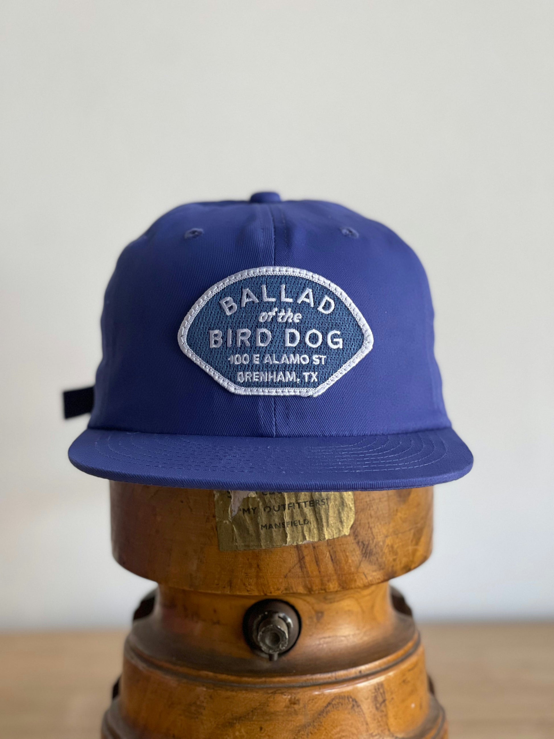 The Shop Hat | Home Base by Ballad of the Bird Dog features a patch with "100 E Alamo St, Brenham, TX," making it the perfect hat for supporting small businesses and your local community. Displayed here on a wooden stand.